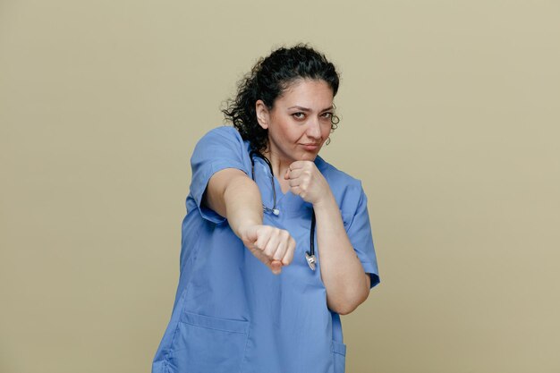 Femme médecin d'âge moyen confiante portant un uniforme et un stéthoscope autour du cou regardant la caméra en gardant le poing sur le menton étirant un autre poing vers la caméra isolée sur fond olive