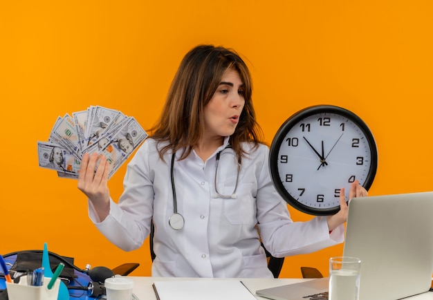 Femme médecin d'âge moyen confus portant une robe médicale avec stéthoscope assis au bureau de travail sur un ordinateur portable avec des outils médicaux tenant une horloge murale et de l'argent sur un mur orange isolé avec espace de copie