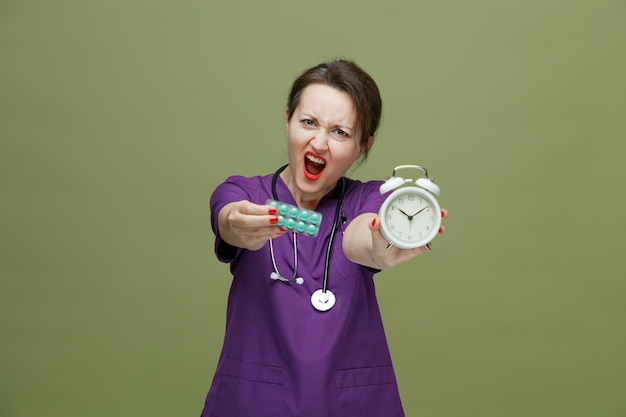 femme médecin d'âge moyen furieuse portant l'uniforme et le stéthoscope autour du cou regardant la caméra étirant le paquet de comprimés et le réveil vers la caméra criant isolé sur fond vert olive