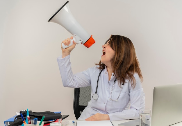 Femme médecin d'âge moyen portant une robe médicale et un stéthoscope assis au bureau avec presse-papiers d'outils médicaux et ordinateur portable à côté criant dans haut-parleur isolé