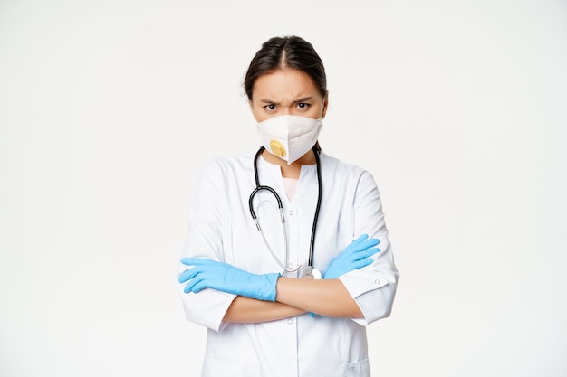 Femme médecin asiatique dans un respirateur médical et des gants en caoutchouc, regardant en colère contre la caméra, les bras croisés sur la poitrine, debout en uniforme de travailleur de la santé, fond blanc.