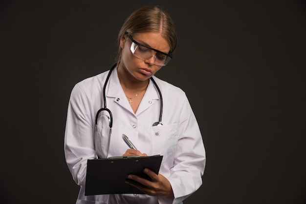 Femme médecin blonde avec un stéthoscope portant des lunettes et en écrivant un reçu au patient.