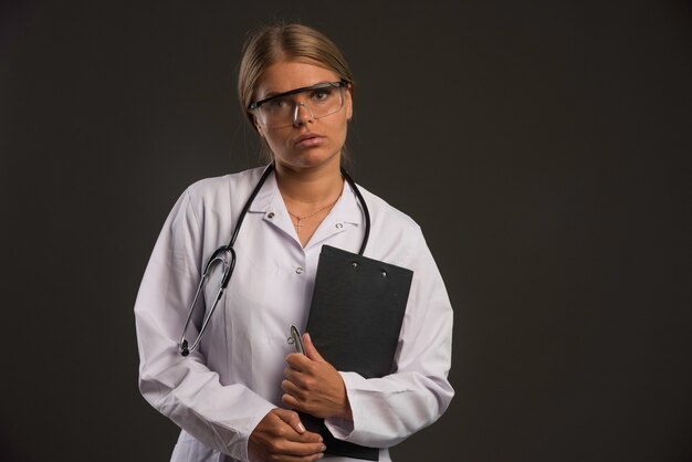 Femme médecin blonde avec un stéthoscope portant des lunettes et tenant un carnet de reçus.