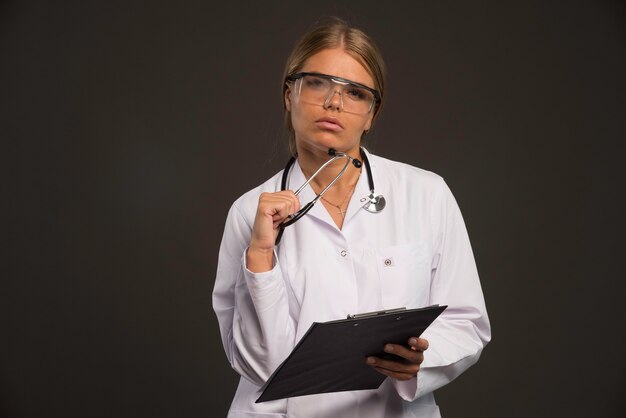 Femme médecin blonde avec un stéthoscope portant des lunettes et tenant un carnet de reçus.