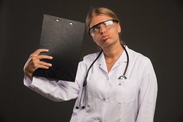 Femme médecin blonde avec un stéthoscope portant des lunettes et touchant la tête avec un carnet de reçus.