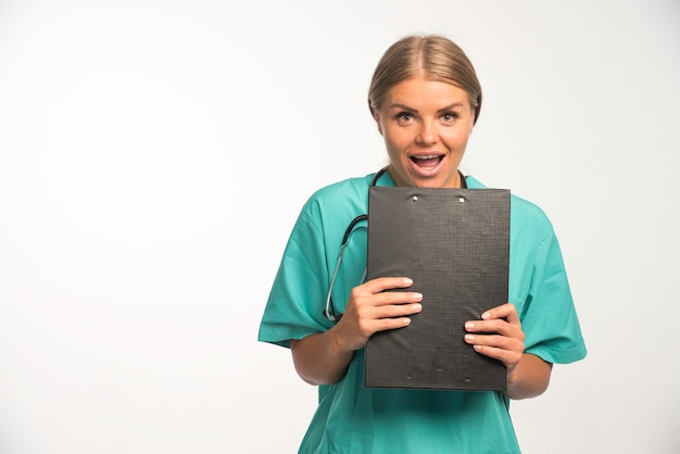 Femme médecin blonde en uniforme bleu tenant un livre de reçus et a l'air excité.