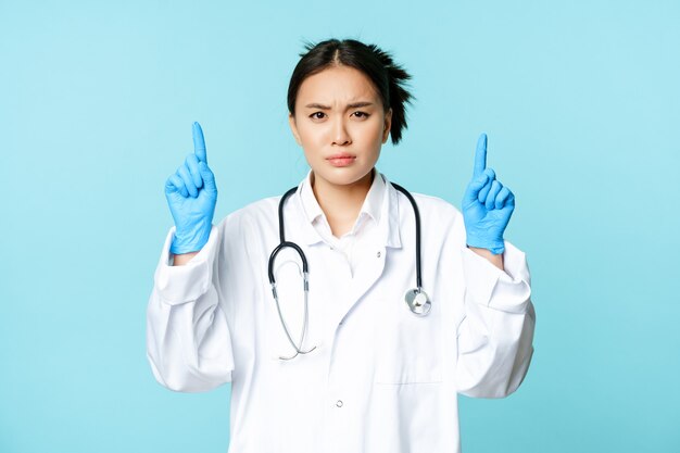 Femme médecin en colère et inquiète, médecin pointant les doigts vers le haut, fronçant les sourcils déçu, debout en uniforme médical sur fond bleu.