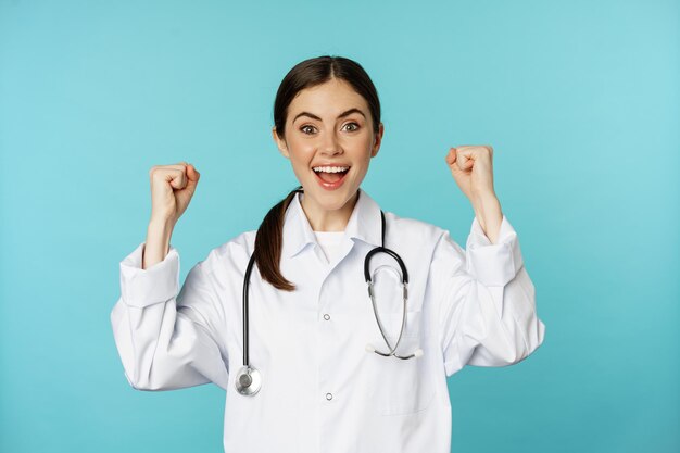 Femme médecin enthousiaste, se réjouissant, gagnant et célébrant, atteindre son objectif, debout en blouse blanche sur fond bleu