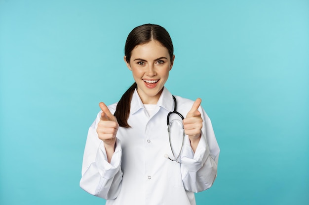 Femme médecin enthousiaste souriant pointant du doigt la caméra félicitant les patients invitant à cli...