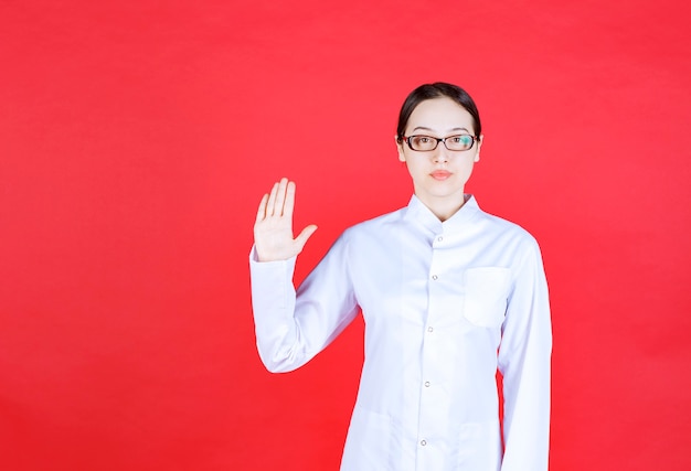 Photo gratuite femme médecin à lunettes debout sur fond rouge et arrêtant quelque chose avec des gestes de la main.