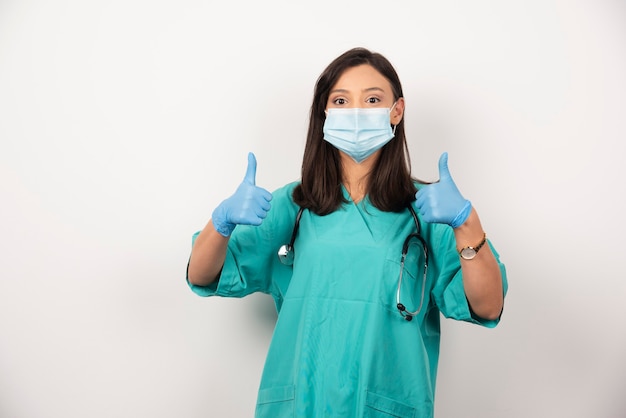 Femme médecin en masque montrant les pouces vers le haut sur fond blanc. Photo de haute qualité