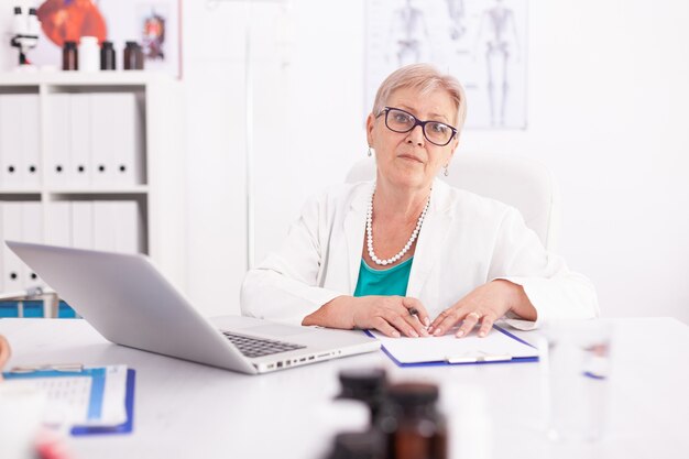 Femme médecin mature portant une blouse de laboratoire dans la chambre d'hôpital lors de l'utilisation d'un ordinateur portable. Médecin utilisant un ordinateur portable sur le lieu de travail de la clinique, confiant, expertise, médecine.