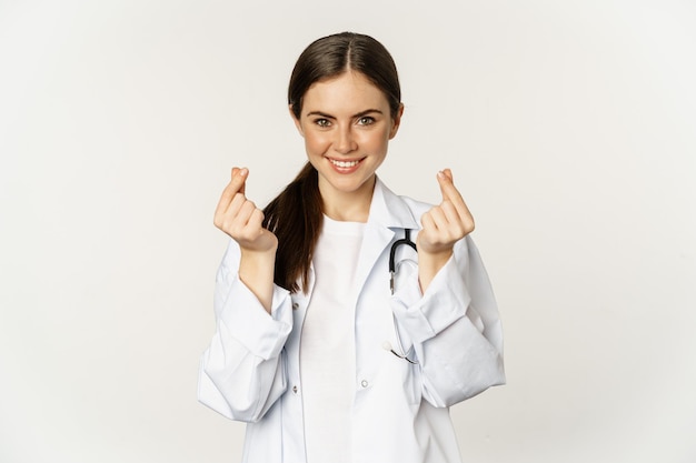 Femme médecin montrant des coeurs de doigt souriant avec soin debout en uniforme sur fond blanc