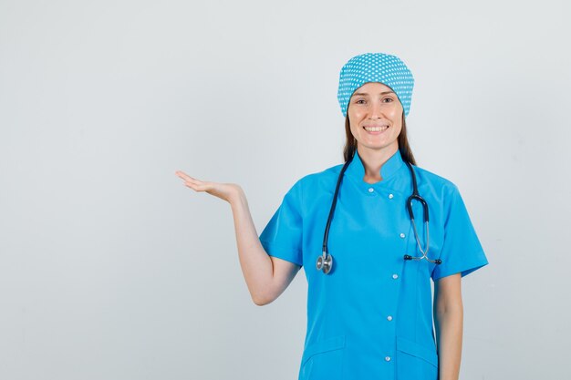 Femme médecin montrant quelque chose avec la main et souriant en vue de face uniforme bleu.