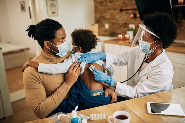 Femme médecin noire examinant un petit garçon avec un stéthoscope lors d'une visite à domicile en raison de la pandémie de COVID19