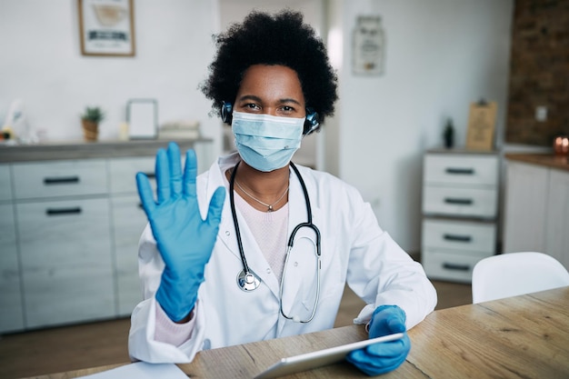 Femme médecin noire avec masque protecteur et gant utilisant un pavé tactile et agitant la caméra