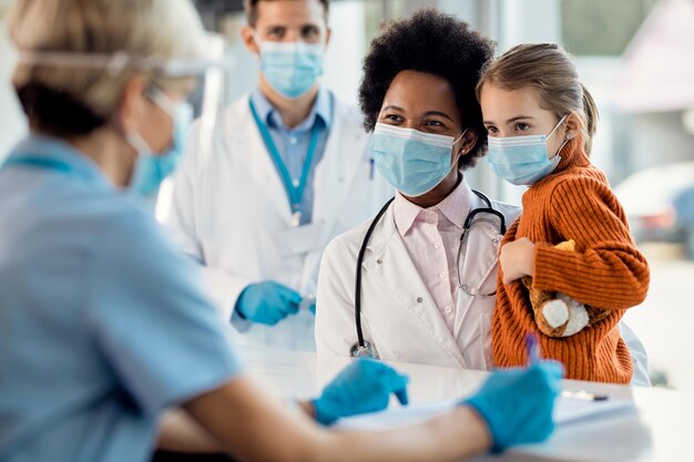 Femme médecin noire et petite fille avec des masques faciaux parlant à une infirmière à la réception