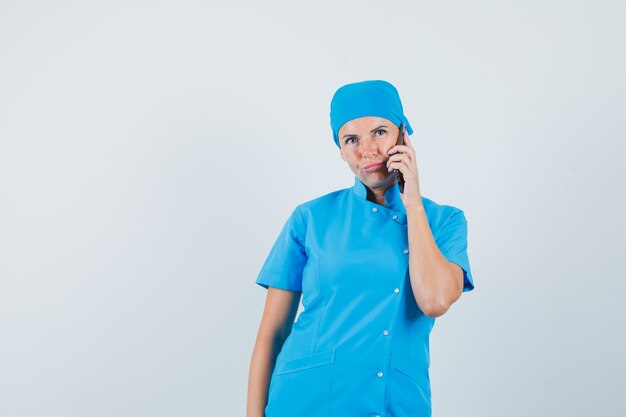 Femme médecin parlant au téléphone mobile en uniforme bleu et à la vue de face, hésitante.