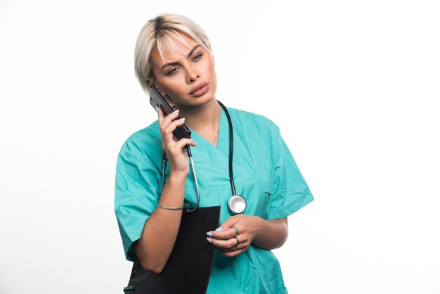 Femme médecin parlant avec téléphone tout en tenant un presse-papiers sur une surface blanche