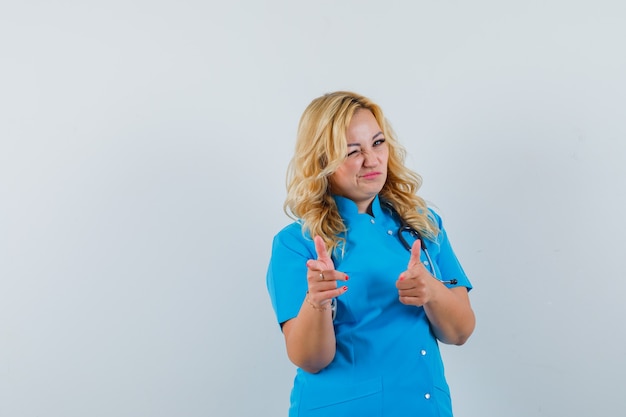 Femme médecin pointant la caméra tout en clignant de l'œil en uniforme bleu et à la recherche assurée. espace pour le texte