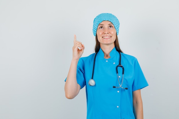 Femme médecin pointant le doigt vers le haut en uniforme bleu et l'air heureux vue de face.
