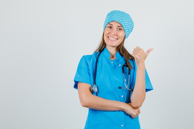 Femme médecin pointant le pouce vers le côté en uniforme bleu et à la joyeuse. vue de face.