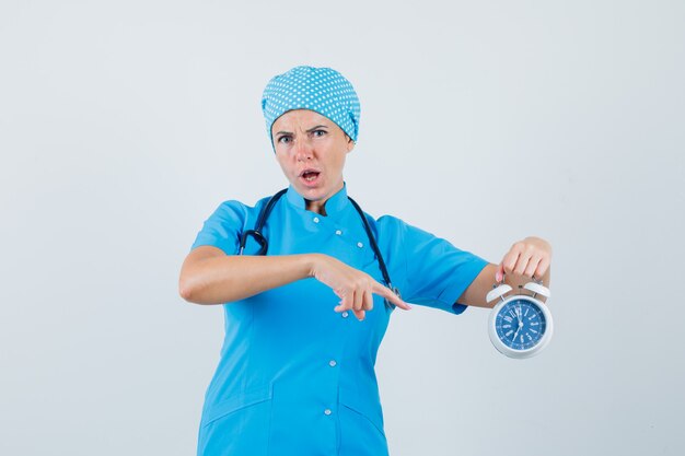 Femme médecin pointant sur réveil en uniforme bleu et à la recherche d'anxiété. vue de face.