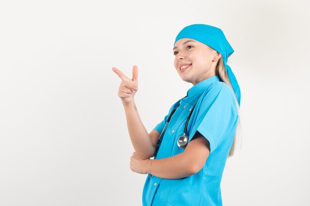 Femme médecin pointant vers le côté avec le geste du pistolet en uniforme bleu et à la bonne humeur.