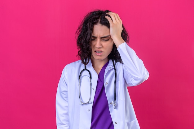 Femme médecin portant blouse blanche avec stéthoscope à la fatigue et onworked debout avec la main sur la tête ayant des maux de tête sur rose isolé