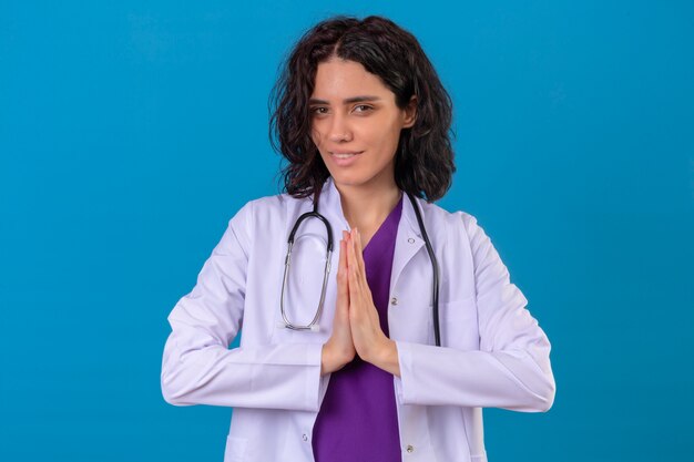 Femme médecin portant une blouse blanche avec stéthoscope, main dans la main dans la prière geste namaste sentiment reconnaissant et heureux sur bleu isolé