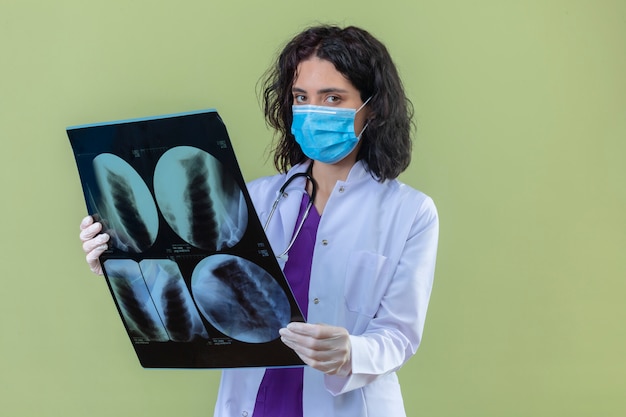 Femme médecin portant blouse blanche avec stéthoscope en masque de protection médicale debout avec x-ray des poumons avec un visage sérieux sur vert isolé