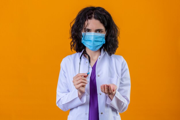 Femme médecin portant blouse blanche avec stéthoscope en masque de protection médicale tenant des pilules dans les mains debout sur orange isolé