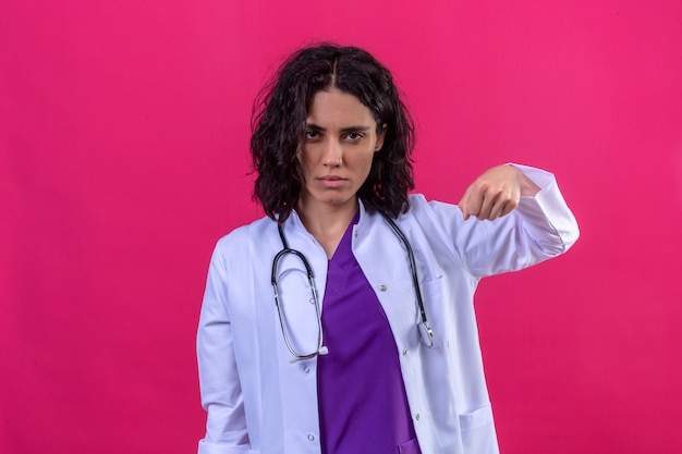 Femme médecin portant une blouse blanche avec stéthoscope montrant le poing à la caméra avec froncement de sourcils menaçant sur rose isolé