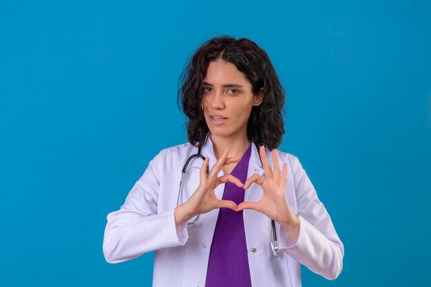 Femme médecin portant blouse blanche avec stéthoscope montrant le symbole du cœur et la forme avec les mains debout sur bleu isolé