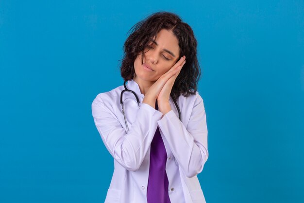 Femme médecin portant une blouse blanche avec stéthoscope à la pose de travail avec les mains ensemble tout en souriant avec les yeux fermés veut dormir sur bleu isolé