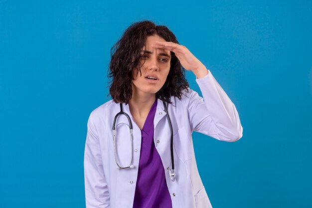 Femme médecin portant blouse blanche avec stéthoscope à la recherche de loin avec la main pour regarder quelque chose debout sur bleu isolé