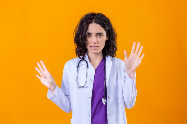 Femme médecin portant blouse blanche avec stéthoscope à la recherche de stressé debout avec les mains levées sur orange