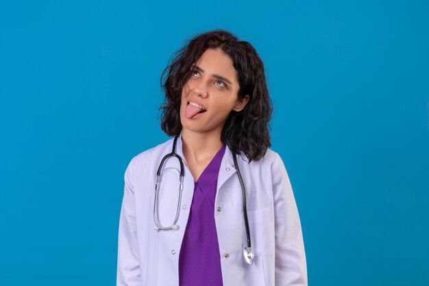 Femme médecin portant blouse blanche avec stéthoscope s'amusant à la joyeuse langue collante à la recherche debout sur bleu isolé