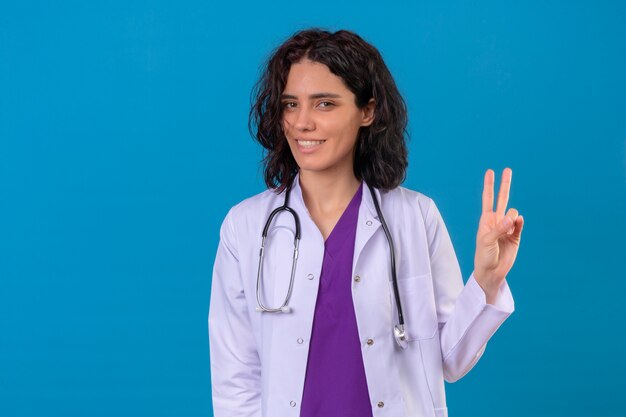 Femme médecin portant blouse blanche avec stéthoscope souriant joyeux à côté et montrant le numéro deux ou signe de la victoire avec les doigts sur bleu isolé