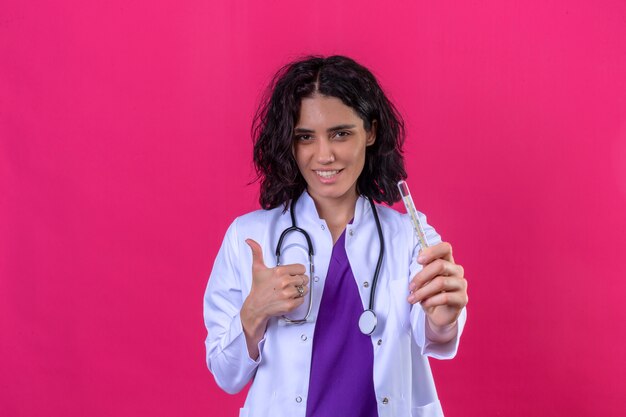 Femme médecin portant blouse blanche avec stéthoscope tenant un thermomètre dans la main montrant le pouce vers le haut avec un visage heureux debout sur rose isolé