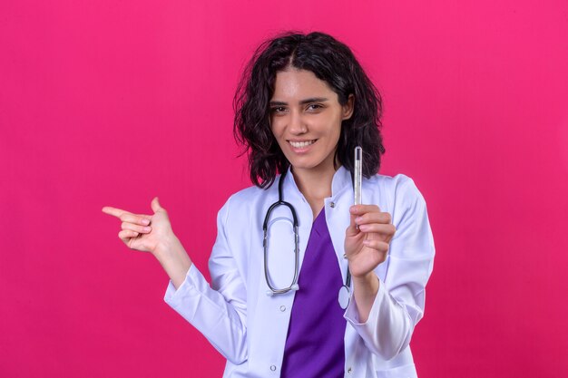Femme médecin portant blouse blanche avec stéthoscope tenant le thermomètre dans la main et pointant avec le doigt sur le côté avec happy face sur rose isolé