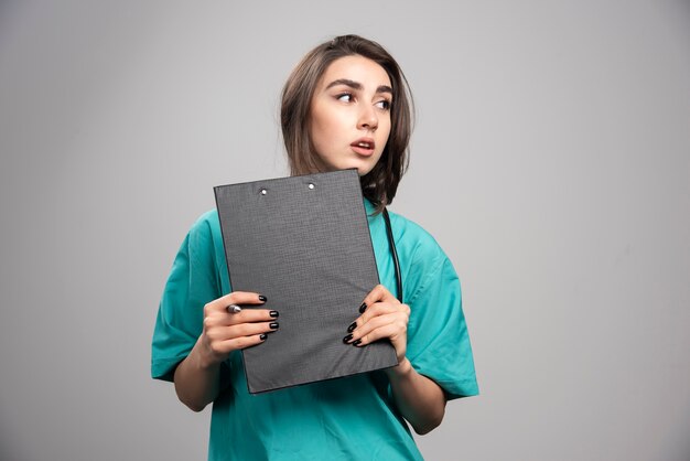 Femme médecin posant avec presse-papiers sur mur gris.