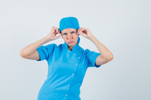 Femme médecin posant tout en se tenant la main à la tête en uniforme bleu et à la pensif. vue de face.