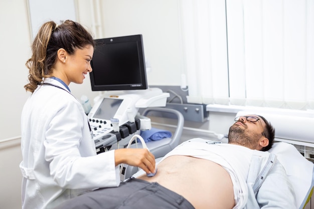 Femme médecin prudente en blouse blanche assise devant un appareil à ultrasons et effectuant des diagnostics abdominaux avec transducteur