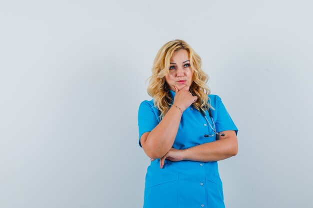 Femme médecin à la recherche de suite avec la main sur la mâchoire en uniforme bleu et à la pensif. espace pour le texte