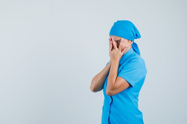 Femme médecin regardant à travers les doigts en uniforme bleu et regardant curieux, vue de face.