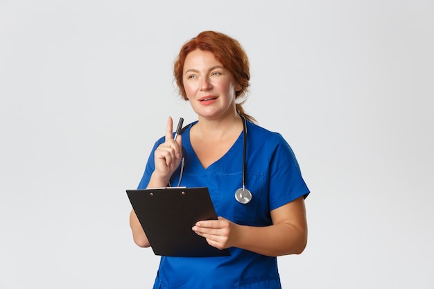 Femme médecin rousse réfléchie, médecin rousse en gommage bleu à la recherche intriguée par le cas du patient, secouant le stylo et tenant le presse-papiers