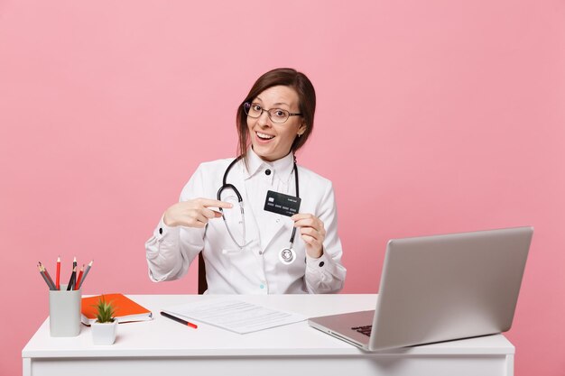Femme médecin s'asseoir au bureau travailler sur ordinateur avec carte de crédit de document médical à l'hôpital isolé sur fond de mur rose pastel. femme en robe médicale lunettes stéthoscope. concept de médecine de santé
