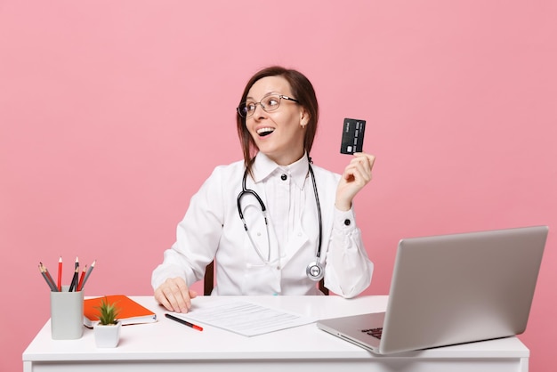 Femme médecin s'asseoir au bureau travailler sur ordinateur avec carte de crédit de document médical à l'hôpital isolé sur fond de mur rose pastel. femme en robe médicale lunettes stéthoscope. concept de médecine de santé