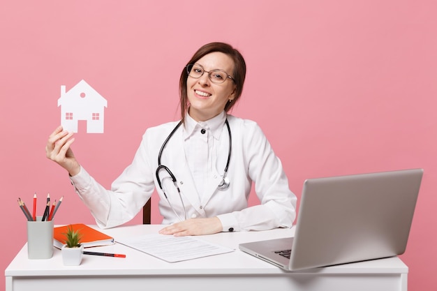Femme médecin s'asseoir au bureau travailler sur ordinateur avec document médical tenir maison à l'hôpital isolé sur fond de mur rose pastel. femme en robe médicale lunettes stéthoscope. concept de médecine de santé.